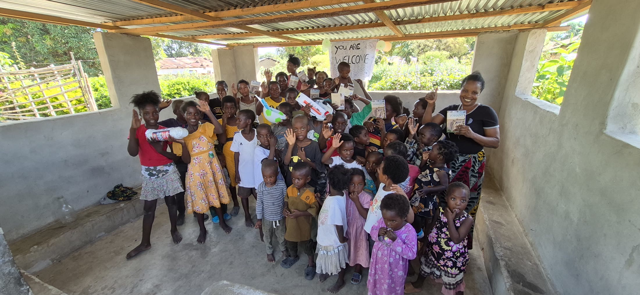 A large group of children and leaders waving inside a village shelter